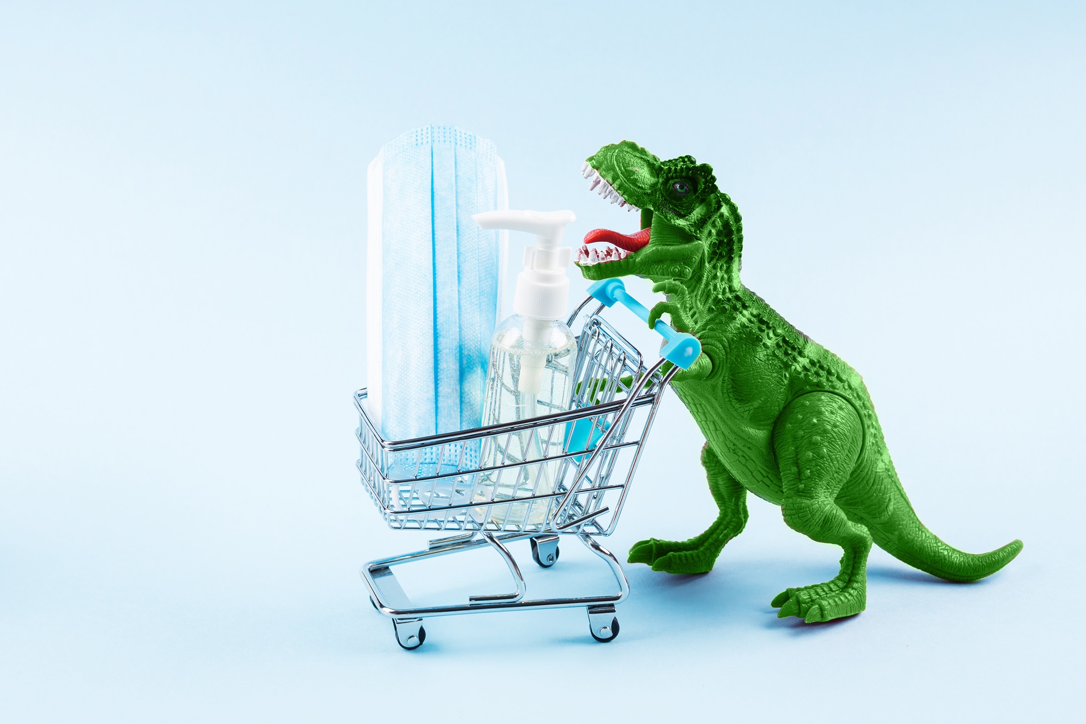 Green toy dinosaur pushing shopping cart containing hand sanitizer and face mask